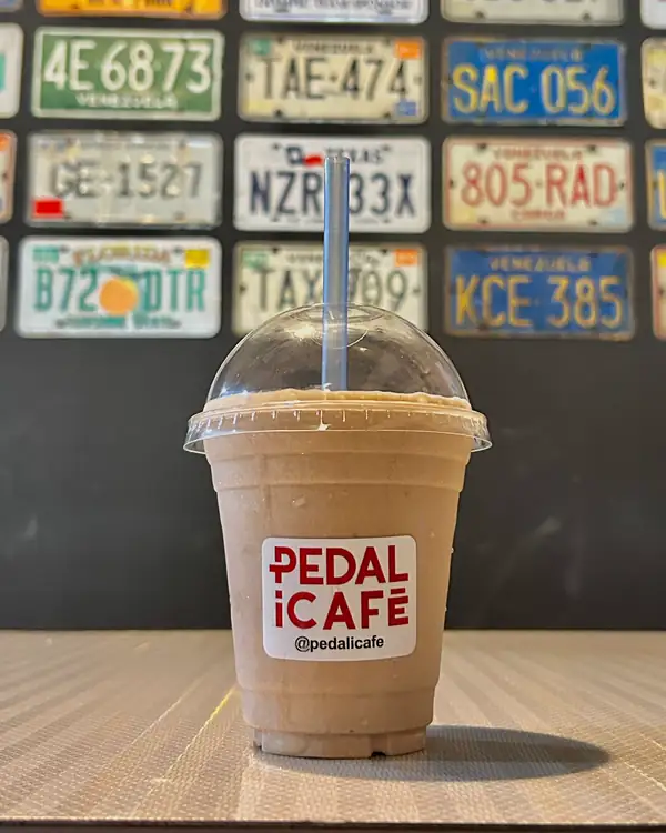 Bebida fría de café en vaso de PEDALiCAFÉ frente a una pared de placas de carros